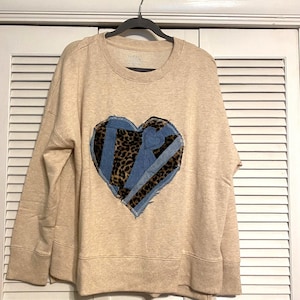 Könnte beinhalten: Beiges Sweatshirt mit großem Herzdesign. Das Herz besteht aus Leopardenmusterstoff und Jeansstoff-Flicken. Das Sweatshirt hat einen Rundhalsausschnitt und lange Ärmel.