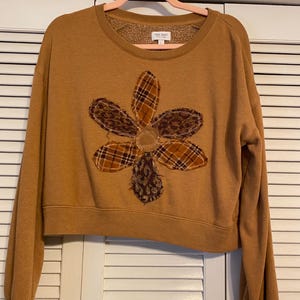 May include: Mustard-colored sweatshirt with a large, appliqued flower design. The flower petals feature plaid and leopard print patterns, with a textured center. The sweatshirt has a crew neck and long sleeves.