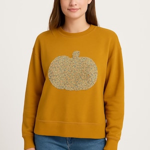 May include: Mustard yellow sweatshirt with a large pumpkin applique. The pumpkin is made of a textured, multi-colored fabric. The sweatshirt has long sleeves and a crew neck. The person is wearing blue jeans.