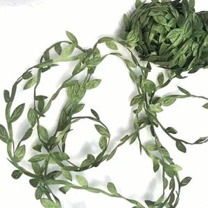 Puede incluir: Guirnalda de hojas verdes artificiales, perfecta para manualidades o decoración. La guirnalda presenta pequeñas hojas detalladas y se muestra enrollada y desenrollada. Las hojas son de un color verde apagado, y los tallos son de un verde más oscuro.
