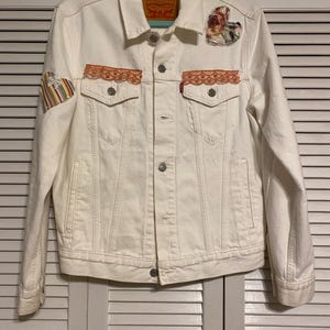May include: White denim jacket with a classic collar, button closure, and two chest pockets. Features decorative patches, including floral and striped designs, and orange lace trim on the pockets. The Levi Strauss & Co. label is visible.