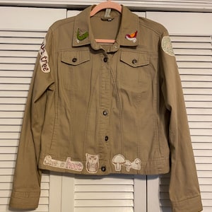 Puede incluir: Chaqueta vaquera color canela con cierre de botones, dos bolsillos en el pecho y parches decorativos. Los parches incluyen un plátano, un pájaro, un búho, setas y el texto "Give a hoot" y "Up the tree".