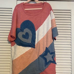 May include: A coral-colored t-shirt with a round neckline and short sleeves. The shirt features a patchwork design with a denim heart and star, along with white and peach-colored stripes. The shirt is hanging on a pink hanger.