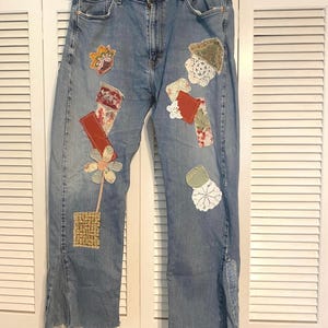 May include: Light blue denim jeans featuring a variety of colorful fabric patches, white lace doilies, and a floral fabric waistband. The jeans have a straight-leg cut with frayed, uneven hems and a vertical slit at the bottom of each leg.