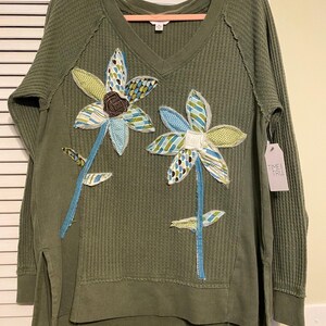 Könnte beinhalten: Olivgrünes Waffelstrick-Langarmshirt mit V-Ausschnitt und Seitenschlitzen. Das Shirt hat zwei große, applizierte Blumendesigns mit blau, grün und weiß gemusterten Blütenblättern und Stielen. Ein Etikett mit der Aufschrift "TIME TRU" ist angebracht.