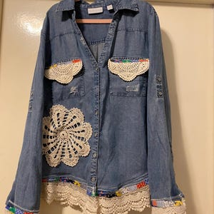 May include: A blue denim shirt with crochet lace details. The shirt features a large crochet doily on the front, crochet lace pockets, and crochet lace trim on the cuffs and hem. The shirt is button-down with a collar.
