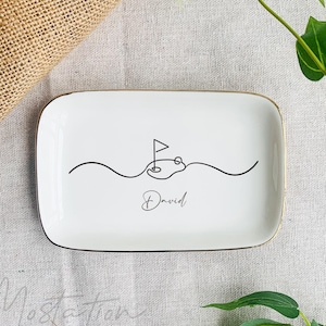 May include: White ceramic trinket tray with a gold rim. The tray features a black line drawing of a golf course with a flag and the word "David" written in cursive.