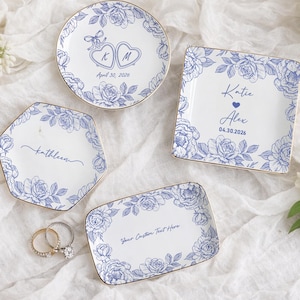 Op de afbeelding: Vier witte keramische ring schaaltjes met blauwe bloemmotieven en gouden randen. Elk schaaltje heeft een andere vorm en gepersonaliseerde tekst, inclusief namen en data. Twee ringen liggen in de buurt van een schaaltje. De schaaltjes liggen op een witte, getextureerde ondergrond.