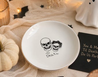 May include: A round white ceramic trinket dish featuring a black line art illustration of two skulls, one wearing a floral crown, with the text 'Til Death' printed below. The dish has a smooth, glossy finish and a simple, circular rim design.