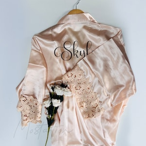 Könnte beinhalten: Blassrosa Satin-Robe mit floraler Spitzenborte an den Ärmeln. Der Name "Skyl" ist in schwarzer Schrift auf der Rückseite aufgedruckt. Ein Holzkleiderbügel und weiße Blumen sind ebenfalls zu sehen.