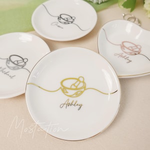以下が含まれることがあります： 金色の縁とパーソナライズされた名前が付いた白いセラミックリング皿。各皿には、金、銀、またはローズゴールドの乳鉢と乳棒のデザインが施されています。「Ashley」、「Michael」、「Maggie」という名前がエレガントな筆記体で表示されています。