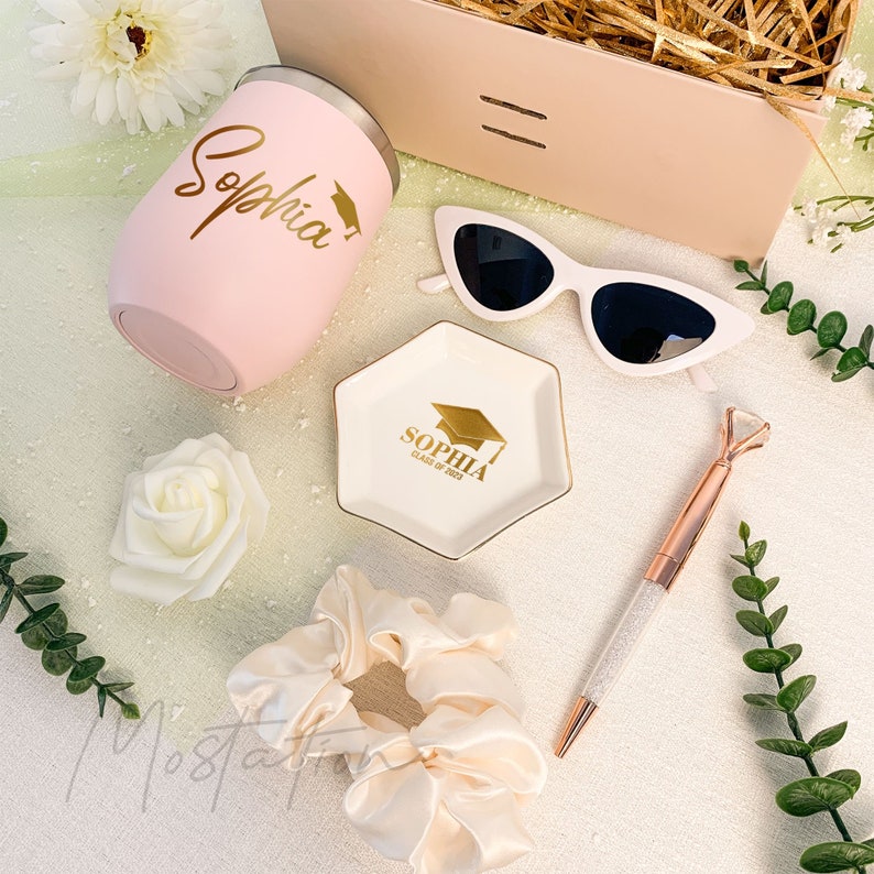 May include: A pink stainless steel tumbler with the name Sophia and a graduation cap in gold, a white hexagonal ceramic trinket dish with gold text Sophia Class of 2023, white cat-eye sunglasses, a rose gold diamond-topped pen, and a cream scrunchie.
