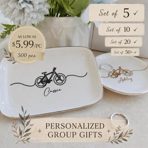 May include: Two white ceramic trinket dishes with gold-toned rims. The rectangular dish features a black line-art bicycle graphic and the name Cassie. The round dish features a gold line-art bicycle graphic and the name Ashley. Text includes Set of 5, 10, 20, 50+, AS LOW AS $5.99/PC 500 pcs, and PERSONALIZED GROUP GIFTS.
