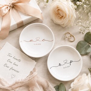 May include: Two round white ceramic ring dishes featuring custom calligraphy. One dish displays the initials N and C with a heart and the date 06.20.2021 in copper. The other dish shows the initials R and P with a heart and the date 06.20.2021 in black.