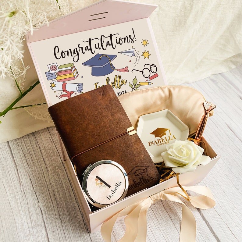 Pu&ograve; includere: Una confezione regalo per la laurea con un diario in pelle marrone, uno specchietto compatto, una mascherina per gli occhi e un piattino. Il coperchio della scatola dice "Congratulazioni!" con un'illustrazione di un cappello di laurea. Il diario e il piatto sono personalizzati con il nome "Isabella".