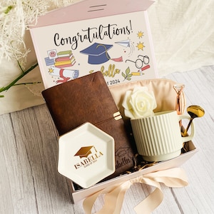 May include: A graduation gift box with a pink lid featuring the text "Congratulations!". Inside are a brown leather journal, a white mug, a small dish with "Isabella Class of 2024" text, a pen, and a decorative rose.