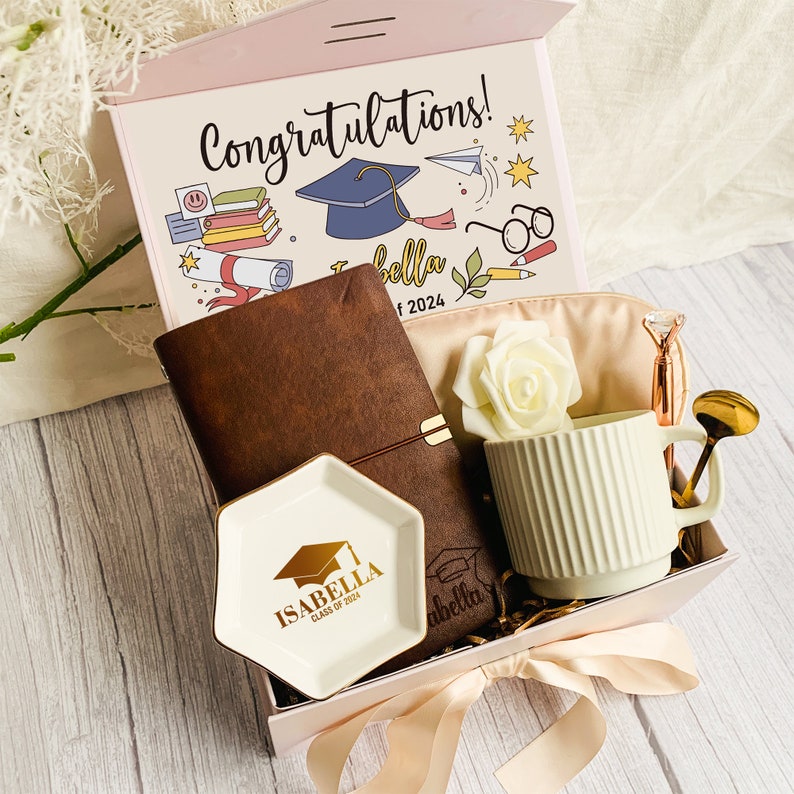 May include: A graduation gift box with a pink lid featuring the text "Congratulations!". Inside are a brown leather journal, a white mug, a small dish with "Isabella Class of 2024" text, a pen, and a decorative rose.