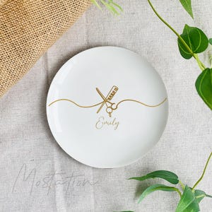 Pu&ograve; includere: Un piattino rotondo in ceramica bianca con una grafica dorata raffigurante forbici da parrucchiere e un pettine incrociati, collegati da una linea ondulata. Il nome Emily &egrave; stampato in corsivo dorato sotto gli strumenti.