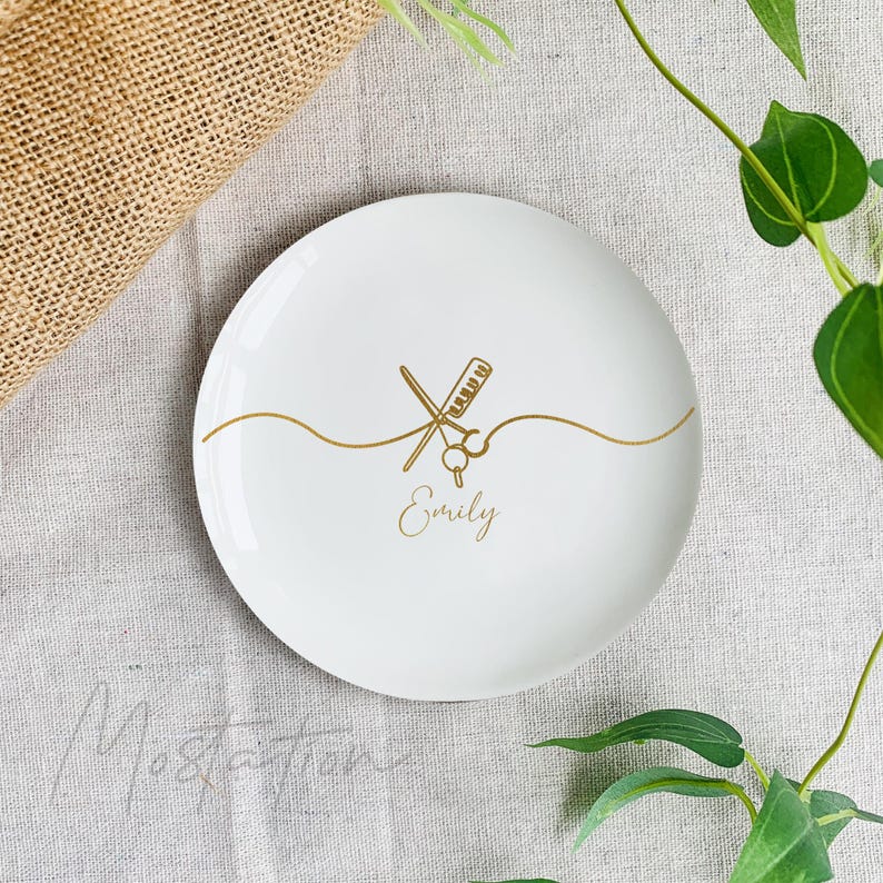 Pu&ograve; includere: Un piattino rotondo in ceramica bianca con una grafica dorata raffigurante forbici da parrucchiere e un pettine incrociati, collegati da una linea ondulata. Il nome Emily &egrave; stampato in corsivo dorato sotto gli strumenti.