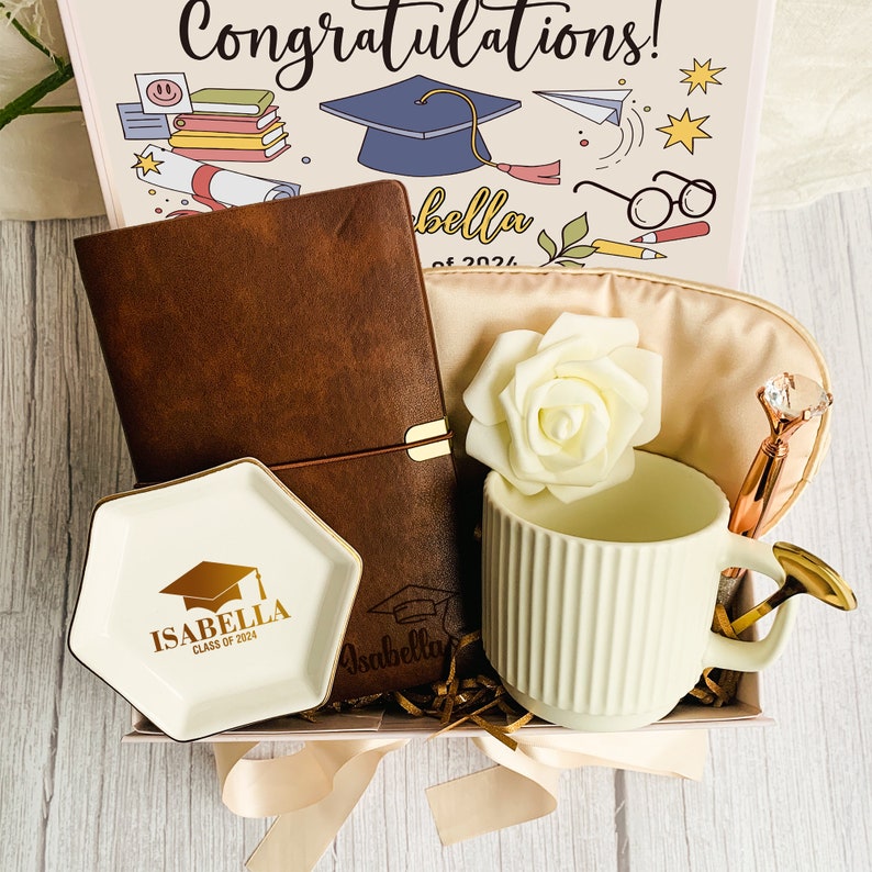 May include: A graduation gift box with a brown leather journal, a white ceramic mug, a small dish, and a sleep mask. The box is decorated with a graduation cap illustration and the text "Congratulations!" and "Isabella Class of 2024".