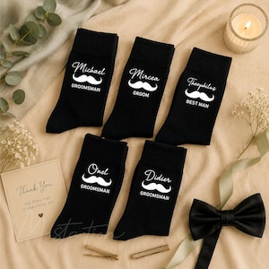 May include: Five black socks featuring white printed text and mustache graphics. The socks are labeled with the names Michael, Mircea, Theophilus, Onel, and Didier, along with titles including Groomsman, Groom, and Best Man, arranged on a fabric surface.