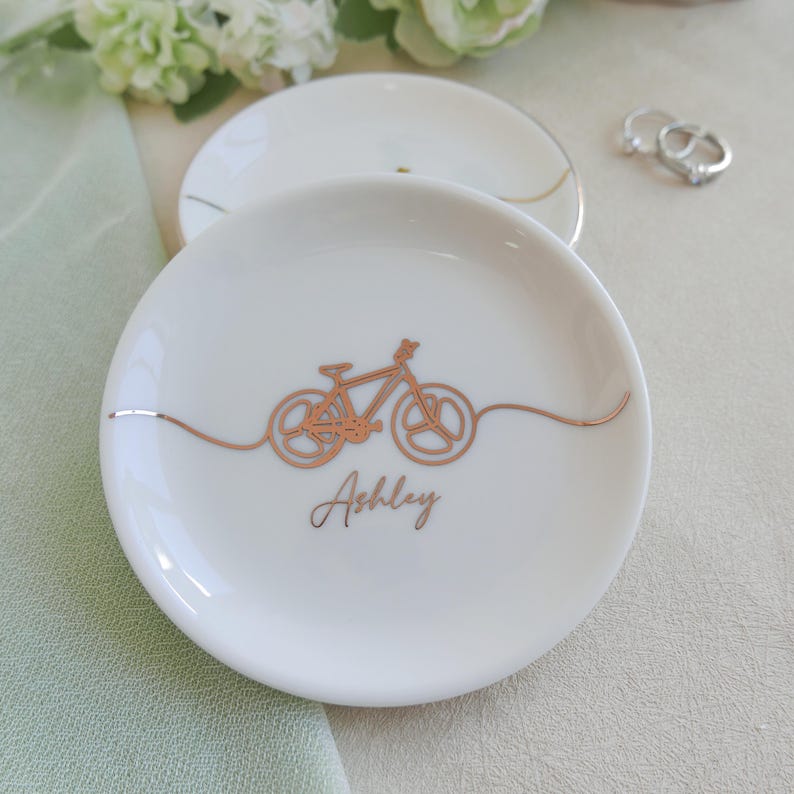May include: A round white ceramic trinket dish featuring a continuous line drawing of a bicycle in metallic copper foil. The name Ashley is printed in a cursive copper font below the bicycle. A thin copper line extends across the dish.