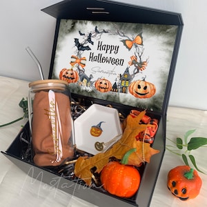 Pu&ograve; includere: Una scatola regalo nera contenente un bicchiere di vetro con coperchio in bamb&ugrave; e cannuccia, un piattino esagonale bianco con illustrazione di zucca, una collana dorata, un panno in tessuto e una zucca finta. Il coperchio presenta una grafica Happy Halloween.