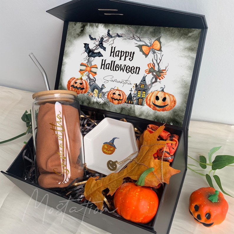 Pu&ograve; includere: Una scatola regalo nera contenente un bicchiere di vetro con coperchio in bamb&ugrave; e cannuccia, un piattino esagonale bianco con illustrazione di zucca, una collana dorata, un panno in tessuto e una zucca finta. Il coperchio presenta una grafica Happy Halloween.