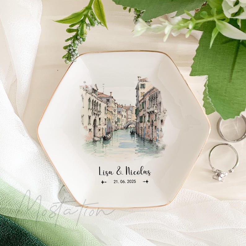 May include: White hexagonal ceramic ring dish with a gold rim. The dish features a watercolor illustration of a canal in Venice, Italy. The text "Lisa & Nicolas 21.06.2025" is printed on the dish.