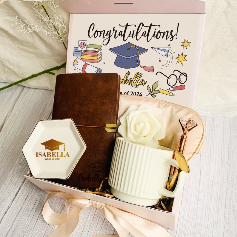 May include: A graduation gift box with a pink lid, containing a brown leather journal, a white mug, a small dish, a pen, and an eye mask. The box lid has the text "Congratulations!" and graduation-themed illustrations.