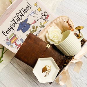 May include: A gift box with graduation-themed items. Includes a journal, mug, eye mask, and a decorative dish. The box lid has the text "Congratulations!" and "Isabella Class of 2024". The items are arranged on a wooden surface.