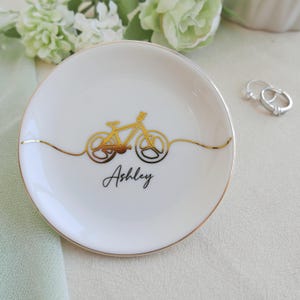 May include: A round white ceramic trinket dish with a gold rim and a gold line drawing of a bicycle centred on the surface. The name Ashley is printed in black script below the bicycle graphic. The dish is positioned near two silver rings.