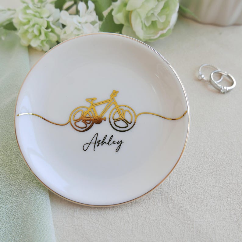 May include: A round white ceramic trinket dish with a gold rim and a gold line drawing of a bicycle centred on the surface. The name Ashley is printed in black script below the bicycle graphic. The dish is positioned near two silver rings.