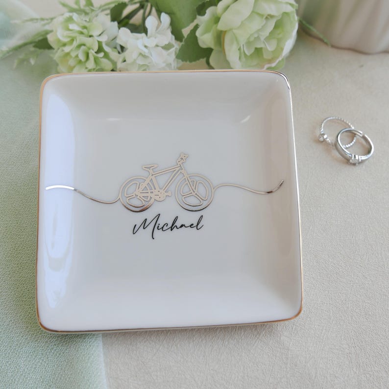 May include: A square white ceramic trinket dish with a gold-trimmed edge. The centre features a metallic silver line-art illustration of a bicycle with a wavy line extending from the wheels. The name Michael is printed in black script below the bike.