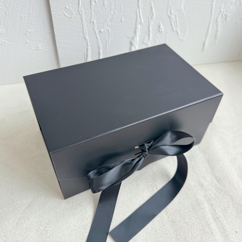 Pu&ograve; includere: Una scatola regalo rettangolare nera con finitura opaca, dotata di coperchio incernierato e un nastro di raso nero abbinato legato a fiocco nella parte anteriore centrale. La scatola presenta bordi netti e una superficie nera liscia e uniforme.