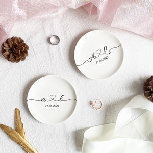 May include: Two round white ceramic ring dishes featuring black cursive lettering with the initials A and L, a heart symbol, and the date 11.06.2022. The dishes are displayed alongside a silver ring, a rose gold ring, pinecones, and ribbon.