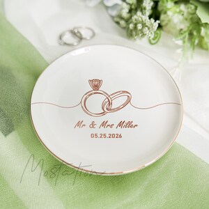 May include: A round white ceramic ring dish with a metallic gold rim. The centre features a copper-coloured line art illustration of two interlocking wedding rings, with the text "Mr & Mrs Miller" and the date "05.25.2026" printed below in copper.