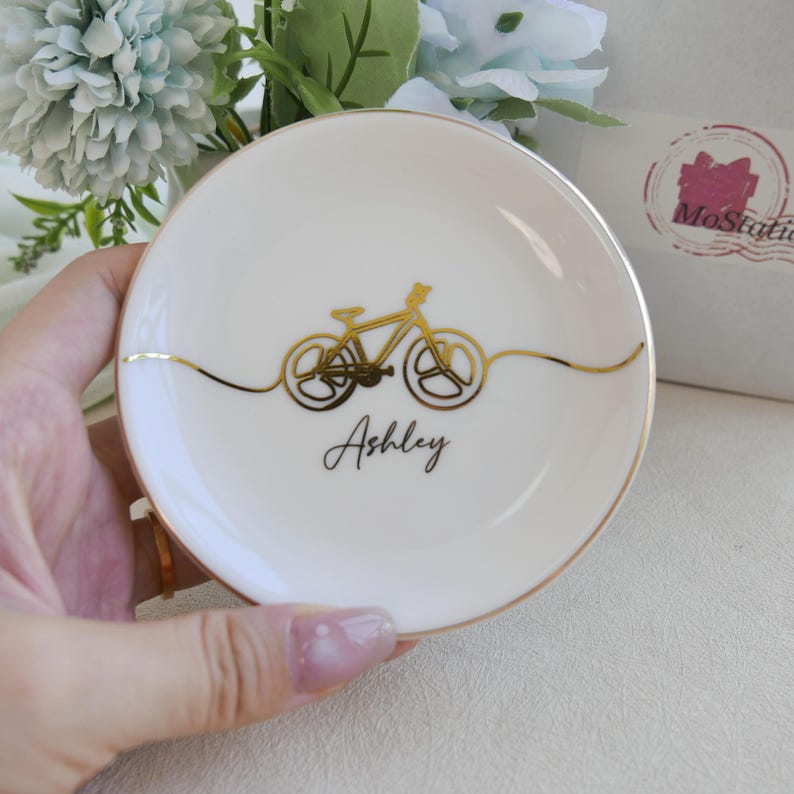 May include: A round white ceramic trinket dish featuring a gold metallic line drawing of a bicycle centred on the surface. Below the bicycle, the name Ashley is printed in a black cursive script. The rim of the dish is finished with a gold edge.