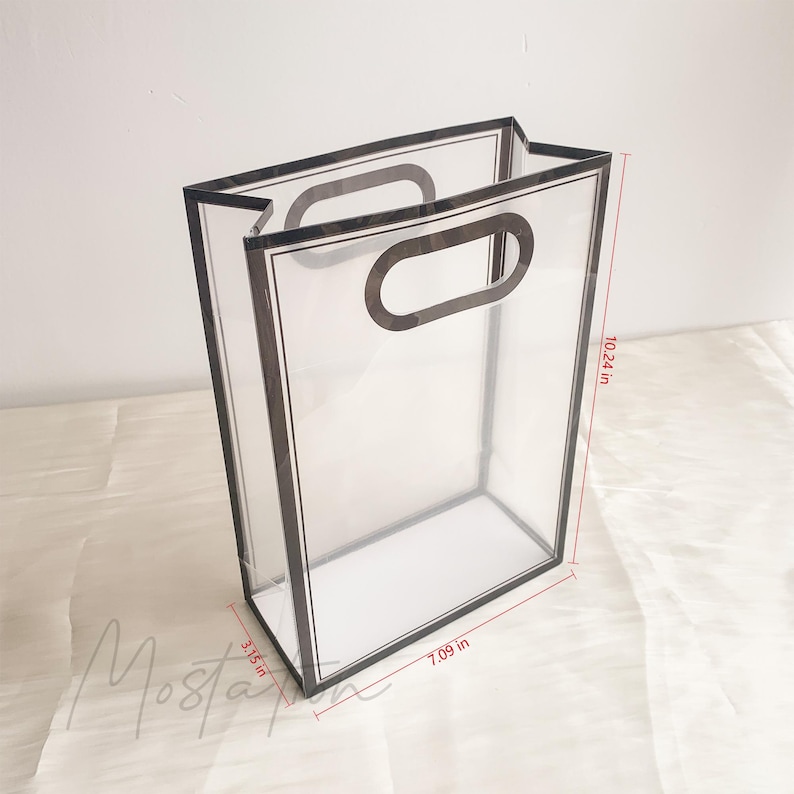 May include: A clear acrylic rectangular gift bag with black trim and integrated oval handles. The bag measures 10.24 inches in height, 7.09 inches in width, and 3.15 inches in depth, as indicated by the red dimension lines on the image.
