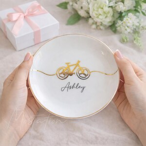 May include: A round white ceramic trinket dish featuring a gold metallic line drawing of a bicycle centred across the middle. Below the bicycle, the name Ashley is printed in a black cursive script. The dish has a thin gold rim around the edge.
