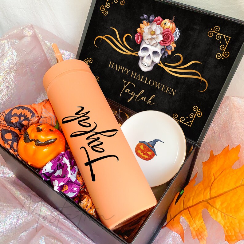 May include: A gift box containing a matte peach tumbler with the name Taylah in black script, a small white ceramic dish with a pumpkin graphic, a miniature orange pumpkin, and patterned fabric scrunchies. The box lid features a skull and floral design.