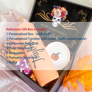 May include: A Halloween gift box containing a personalized peach-colored tumbler with a straw, a white ring dish with a pumpkin illustration, a pumpkin ornament, scrunchies, a twist headband, and raffia. The box lid features a skull and floral design.