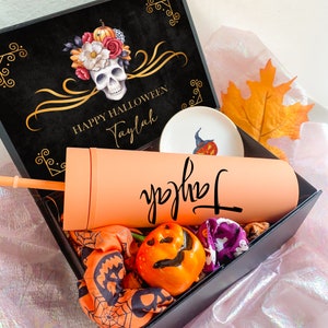 May include: A black gift box containing a peach-colored tumbler with the name Taylah in black script, a matching straw, a small white dish with a pumpkin illustration, a pumpkin-shaped item, and orange and purple patterned fabric accessories.