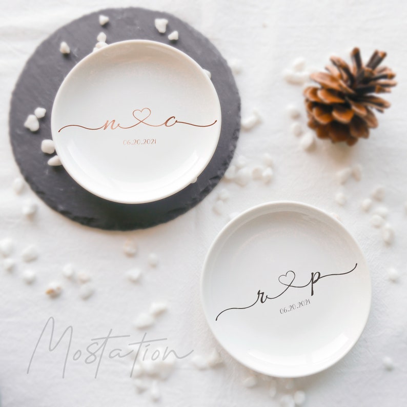 May include: Two round white ceramic trinket dishes featuring personalized lettering. One dish displays the letters n and c in copper with a heart and the date 06.20.2021. The other dish features the letters r and p in black with a heart and the date 06.20.2021.