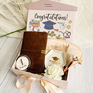 Pu&ograve; includere: Confezione regalo di laurea con coperchio rosa a forma di casa. All'interno, un diario in pelle marrone, uno specchietto compatto, un piattino bianco, una rosa e una penna. Il coperchio recita "Congratulazioni!" con un'illustrazione di cappello di laurea.