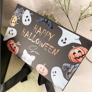 Pu&ograve; includere: Una scatola regalo rettangolare nera con la scritta "HAPPY HALLOWEEN" in arancione e il nome "Sam" scritto sotto. La scatola &egrave; decorata con illustrazioni di fantasmi bianchi, zucche arancioni, un pipistrello nero, un cappello da strega e una ragnatela.