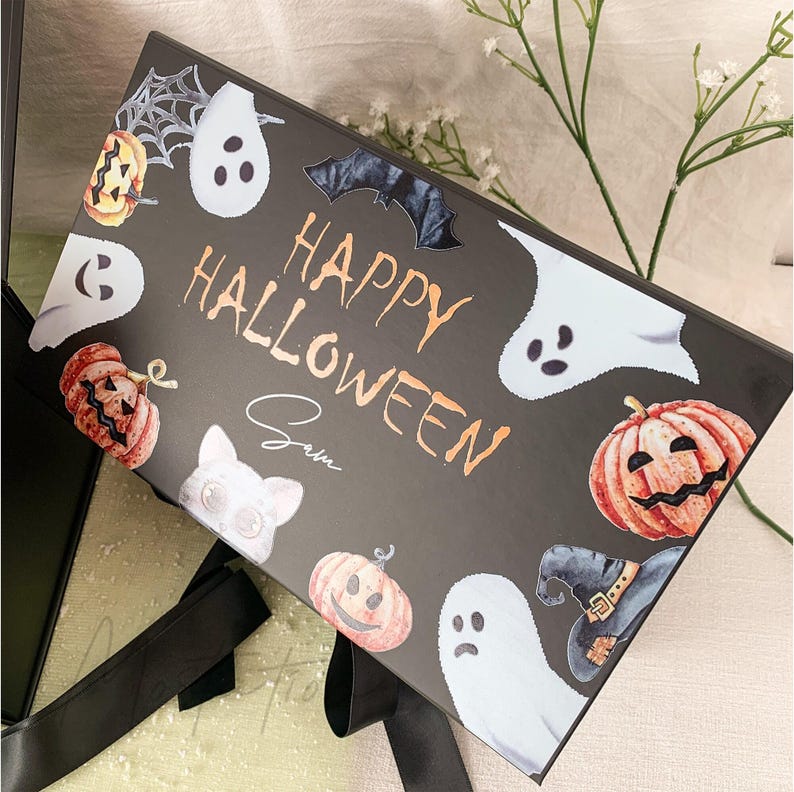 Pu&ograve; includere: Una scatola regalo rettangolare nera con la scritta "HAPPY HALLOWEEN" in arancione e il nome "Sam" scritto sotto. La scatola &egrave; decorata con illustrazioni di fantasmi bianchi, zucche arancioni, un pipistrello nero, un cappello da strega e una ragnatela.