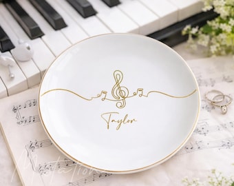 May include: A round white ceramic trinket dish with a gold rim. The centre features a gold line art design of a treble clef and musical notes, with the name Taylor written in gold cursive script below the musical symbols.