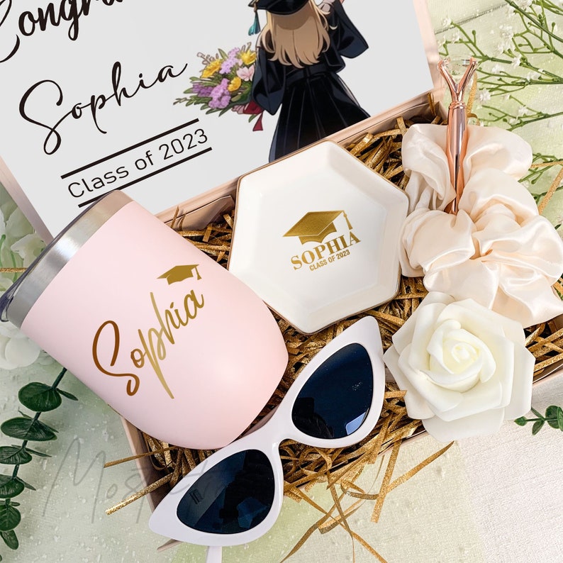 May include: A graduation gift box containing a light pink tumbler with the name Sophia and a graduation cap icon, a white hexagonal trinket dish with gold text, a rose gold pen, two cream-colored satin scrunchies, white sunglasses, and a rose.