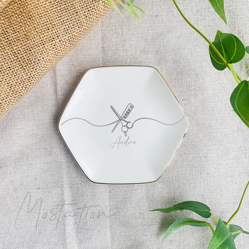 Pu&ograve; includere: Un piattino esagonale in ceramica bianca con bordo dorato. Al centro &egrave; presente un disegno a linee grigie di un pettine e forbici incrociati su una linea ondulata, con il nome Andre stampato in carattere corsivo sotto il disegno.